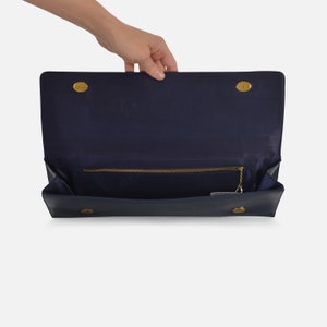 Puede incluir: Bolso de mano azul marino con cierres y herrajes dorados. El interior est&aacute; forrado con tela azul marino y tiene un bolsillo con cremallera. El bolso est&aacute; abierto, revelando el interior y una peque&ntilde;a cadena.