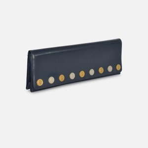 Puede incluir: Una cartera de cuero azul marino con una fila de tachuelas circulares decorativas doradas y plateadas. La cartera es rectangular y parece estar cerrada. Las tachuelas est&aacute;n espaciadas uniformemente en la parte delantera de la cartera.