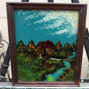 May include: A framed painting of a small cottage with a red roof, surrounded by trees and a stream. The sky is blue with white clouds. The painting is in a dark wood frame.