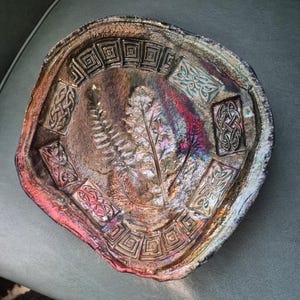 May include: A handcrafted, iridescent ceramic bowl with a unique, organic shape. The bowl features embossed Celtic knot designs and a central fern leaf motif. The color palette includes shades of brown, purple, and pink, creating a captivating visual effect.