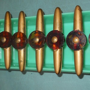 May include: A set of eight vintage cabinet pulls. Each pull has a gold-toned handle and a round, decorative center with a mottled brown and amber pattern. They are arranged in a green rectangular tray.