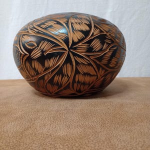 May include: A rounded, bowl-shaped object with a dark black finish and intricate, light brown carved leaf patterns covering the entire exterior surface. The design features repeating, textured lines that radiate from the center of the curved form.