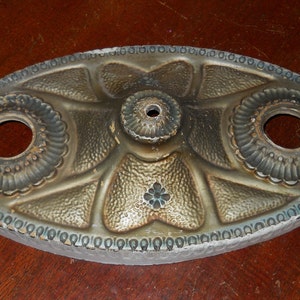 May include: Oval-shaped metal ceiling fixture with a decorative design. The fixture has a central hole and two smaller holes on either side. The metal is a gold color with a textured surface.
