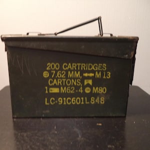 May include: A dark green metal ammunition box with a black handle. The box has yellow text that reads "200 CARTRIDGES 7.62 MM, M 13 CARTONS, 1 M62-4 M80 LC-91C601L848". The box is sitting on a dark surface.