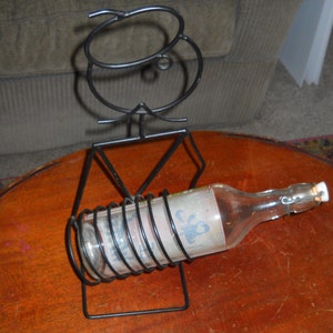 May include: Black metal wine bottle holder shaped like a person with a bottle of wine inside. The bottle has a clear glass body and a white label with a design.
