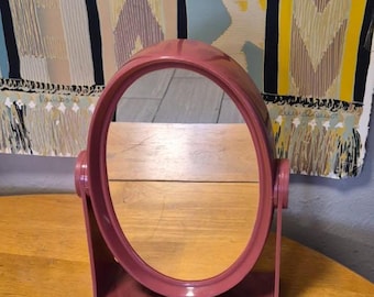 May include: A dusty rose oval-shaped tabletop mirror with a rotating frame. The mirror is mounted on a matching dusty rose plastic stand with a rectangular base, allowing the oval glass to tilt and pivot on two side hinges for adjustable viewing.