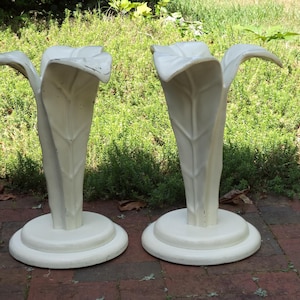 May include: A pair of off-white candle holders featuring a sculptural design that mimics the shape of a large, upright leaf or flower petal. Each holder has a wide, circular base and a textured, organic form with visible veins and edges.