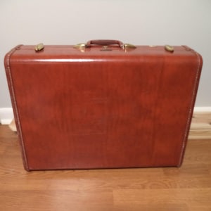 May include: A vintage brown suitcase with brass-colored hardware. The rectangular suitcase has a handle on top and is made of a glossy material. The suitcase is sitting on a wooden floor.