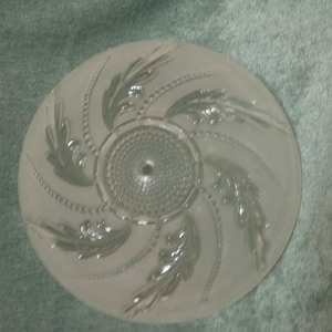 May include: A circular, frosted glass light fixture shade featuring a central medallion with a textured, beaded pattern. Four embossed, swirling leaf motifs extend outward from the center, interspersed with delicate, curved beaded lines.