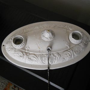 May include: White porcelain ceiling light fixture with three sockets and a decorative chain. The fixture has a floral design and is in the shape of an oval.