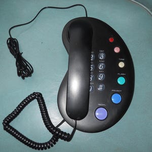 May include: Black, oval-shaped telephone with a handset and a coiled cord. The phone has a keypad with numbers and function buttons, including "TONE", "FLASH", and "PRIVACY". Various colored buttons and indicator lights are visible.
