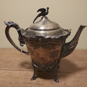 May include: Silver teapot with a pheasant finial on the lid. The teapot has an ornate design with a floral motif and three legs.