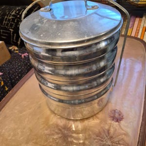 May include: A multi-tiered, cylindrical metal food container featuring four stacked compartments with a secure lid. The container is constructed from polished silver-toned metal and includes a sturdy, arched metal handle attached to the sides for carrying.