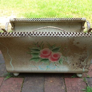 May include: Vintage metal planter with a distressed finish. The planter features a floral design with pink roses and green leaves. It has a decorative, openwork trim and a handle. The planter is supported by small, round feet.