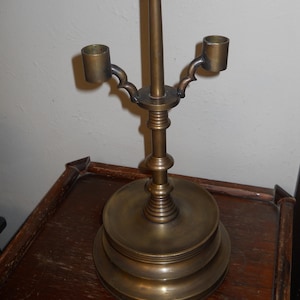 May include: A brass candlestick holder with two arms and a central spike. The base is round and tiered, and the arms have decorative curves. The candlestick is a warm, antique gold color.