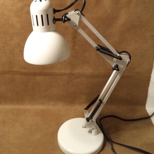 May include: A white desk lamp with an adjustable arm and a round base. The lamp has a white shade with vertical slits and a black power cord. The lamp's arm is articulated, allowing for flexible positioning. The base is approximately 6 inches in diameter.