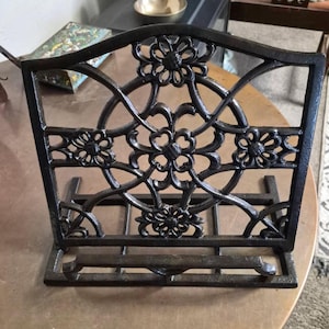 May include: A black cast iron book stand with a floral design. The stand features an arched top and a series of decorative flowers and swirling patterns. The base has a ledge to hold the book open.