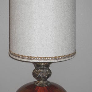May include: A vintage table lamp with a white textured lampshade and a decorative gold trim. The base features a round, orange-red glass globe supported by ornate silver-toned metalwork. The lamp is a classic piece of home decor.