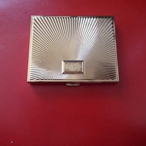May include: A gold-colored metal compact with a sunburst design and a rectangular inscription in the center.