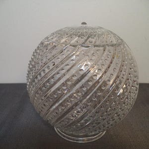May include: A clear, spherical glass jar with a lid. The jar has a spiral pattern of raised dots and lines. The lid has a small knob on top. The jar sits on a clear, circular base. The jar is likely used for storage or display.