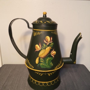 May include: Dark green metal teapot with a black handle and spout. The teapot is decorated with a yellow bird and floral design. The teapot has a lid with a small yellow knob and yellow trim.