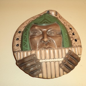 Puede incluir: Un adorno de pared de cerámica que representa a una persona tocando una flauta de pan. La persona tiene la cara marrón con el pelo verde y marrón y lleva un tocado verde y marrón. La flauta de pan es marrón y tiene una raya blanca.