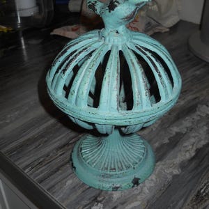 May include: A distressed teal-colored decorative birdcage with a bird perched on top. The cage has a round, open design with vertical bars and sits on a pedestal base. The metal has a weathered appearance with dark brown accents.