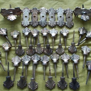 May include: A collection of 25 silver-toned metal decorative screws with ornate floral designs. The screws are arranged in rows on a green surface.