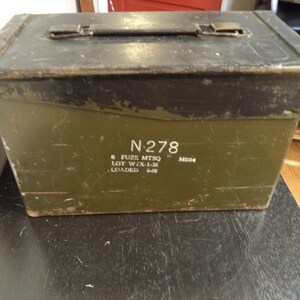 May include: An olive green metal box with a hinged lid and a handle. The box has the text "N 278" and other markings. The top of the box is dark, and the box appears to be aged.