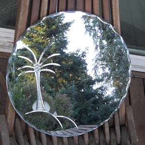 May include: A round, clear glass mirror with a decorative etched palm tree design. The mirror has a scalloped edge and reflects a view of green trees and foliage. The mirror is mounted on a wooden surface.