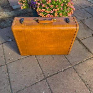 May include: A rectangular, tan-colored hard-shell suitcase featuring a central handle, two metallic latches on the top edge, and subtle horizontal line detailing. The suitcase has a smooth, worn finish and is positioned upright on a paved surface.