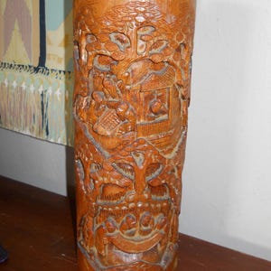 May include: A carved wooden vase with a brown finish. The vase is decorated with a detailed scene of people and trees.