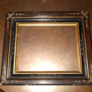 May include: Rectangular picture frame with a black and gold design. The frame has a black outer border with gold accents and a gold inner frame. The corners feature decorative arrow and leaf patterns. The frame is empty.