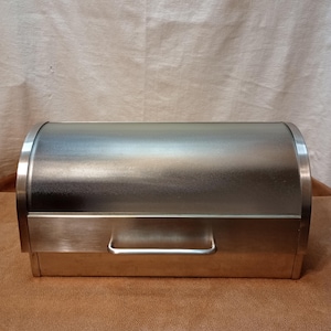 May include: A rectangular bread box with a brushed stainless steel finish and a curved, roll-top lid. The front features a horizontal metal handle attached to the lower section, providing access to the interior storage space for baked goods.