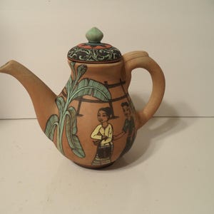 May include: A terracotta teapot featuring a hand-painted scene of two figures standing near a banana tree. The teapot has a curved handle, a tapered spout, and a matching lid with a small green knob. The surface displays earthy brown tones.