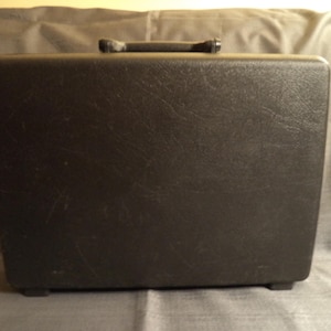 May include: A black, rectangular carrying case with a textured surface. The case has a handle on top and small feet on the bottom. The case is likely for a typewriter or other small equipment.