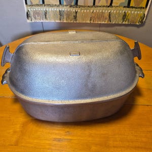 May include: A vintage, oval-shaped, cast-iron Dutch oven with a textured, silver-colored exterior. The oven has a lid and handles on each side. It sits on a wooden table, suggesting it's ready for cooking.