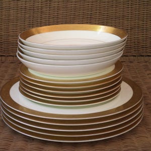 May include: A stack of white ceramic plates and bowls with gold rims. The plates are stacked on top of each other, creating a tiered effect. The gold rims add a touch of elegance to the tableware.
