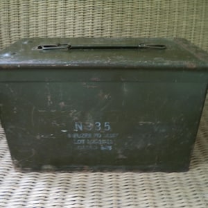 May include: An olive green metal box with a hinged lid and a handle. The box has visible wear and tear, with faded markings including "N 935" and other text. The box is rectangular and sits on a woven surface.