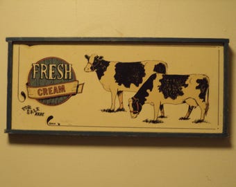 Könnte beinhalten: Ein Holzschild mit einem blauen Rahmen und cremefarbenem Hintergrund. Das Schild zeigt eine Schwarzweißillustration von zwei Kühen und den Text "FRESH CREAM" in einem roten und blauen Kreis.