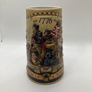 May include: Tan ceramic beer stein with a detailed relief depicting historical figures and an American flag. The stein features the text "1776" and "Birth of a Nation", with the Miller logo and dates 1855 and 1993.