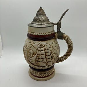 May include: A ceramic beer stein with a pewter lid and a rope handle. The stein features a detailed image of a sailing ship with the text "Bark T. Staysail" on the bottom.