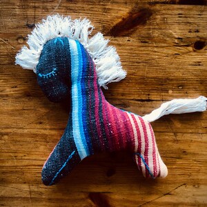Op de afbeelding: Een handgemaakt stoffen paardenspeelgoed met een kleurrijk gestreept lichaam in blauw, rood en wit. Het paard heeft een witte franje manen en staart, en geborduurde gesloten ogen. Geplaatst op een houten oppervlak.