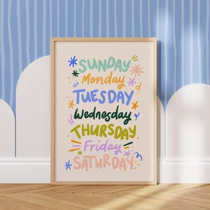 May include: A framed print featuring the days of the week in colorful, whimsical lettering. The days are arranged vertically, with each day highlighted with decorative elements like stars and lines. The frame is a light wood color, and the background is a light beige.
