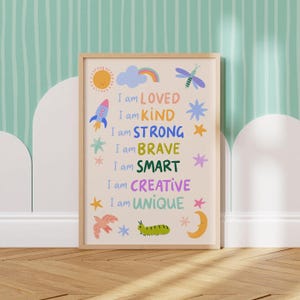 May include: A framed art print with a light wood frame. The print features colorful text and illustrations, including a sun, rocket, and stars. The text reads "I am LOVED, KIND, STRONG, BRAVE, SMART, CREATIVE, UNIQUE."