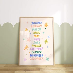 May include: A framed print featuring the months of the year in colorful text. Each month is written in a different color, with decorative starbursts. The background is a light beige color, and the frame is a light brown.