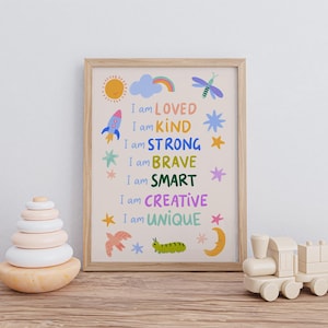 May include: A framed print with the words "I am LOVED, KIND, STRONG, BRAVE, SMART, CREATIVE, UNIQUE" in colorful text. The print features illustrations of a sun, rocket, cloud, and other whimsical designs. A wooden toy train and stacking rings are also visible.
