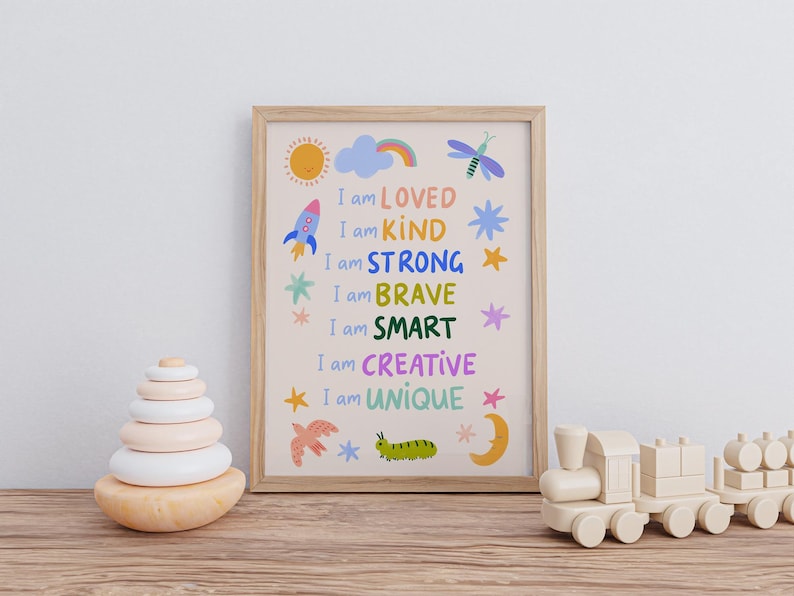 May include: A framed print with the words "I am LOVED, KIND, STRONG, BRAVE, SMART, CREATIVE, UNIQUE" in colorful text. The print features illustrations of a sun, rocket, cloud, and other whimsical designs. A wooden toy train and stacking rings are also visible.