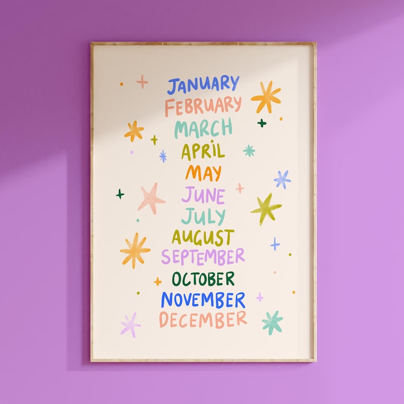 May include: A framed art print on a cream background with the months of the year in colorful, handwritten text. Each month is accompanied by a star or plus symbol. The frame is a light brown color, and the background is a solid purple.