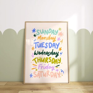 May include: A framed art print featuring the days of the week in colorful, whimsical lettering. The words are in various colors, including blue, green, yellow, and pink, with decorative star and line accents. The frame is a light wood color.